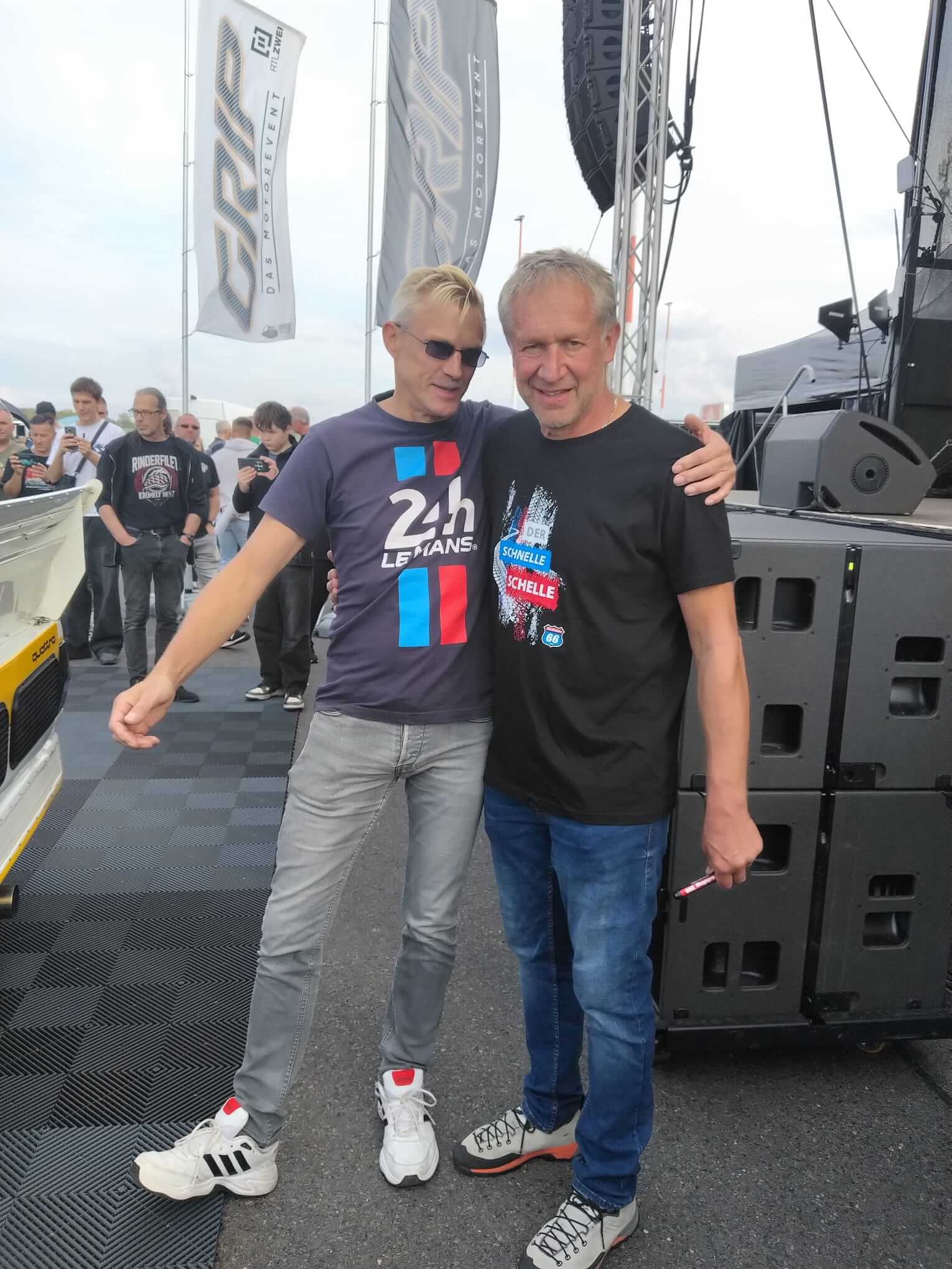 Zwei Männer umarmen sich vor einer Bühne bei einer Motorsportveranstaltung.